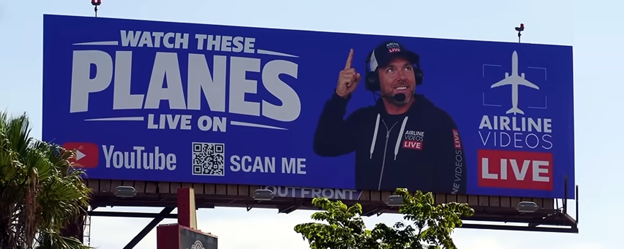 The installed Airline Videos Live billboard in Los Angeles — Watch These Planes Live On YouTube headline, QR code, the channel host pointing skyward, and the Airline Videos Live logo on a royal blue field, mounted on an OUTFRONT structure