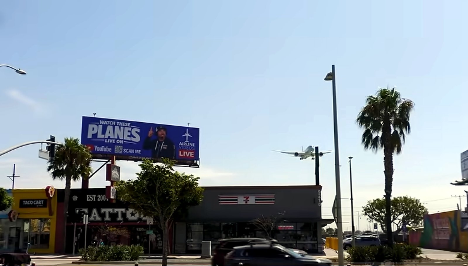The Airline Videos Live billboard above a Los Angeles street, with an airliner on final approach passing to its right