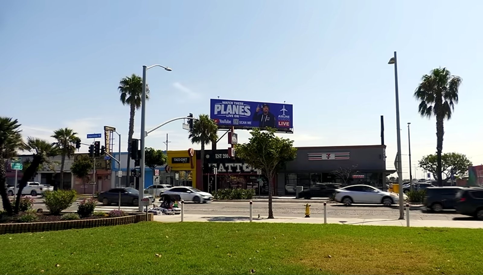 Street-level view of the Airline Videos Live billboard above a row of storefronts, palm trees, and traffic in Los Angeles