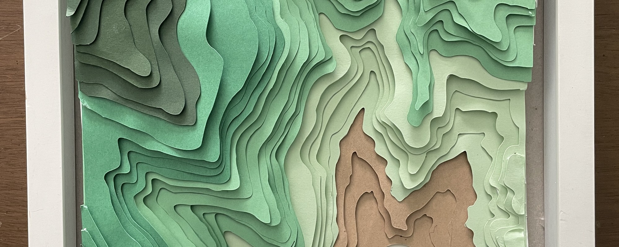 The finished layered-paper topographic shadowbox, framed — stacked cardstock contours in graduated greens stepping down to a tan valley floor